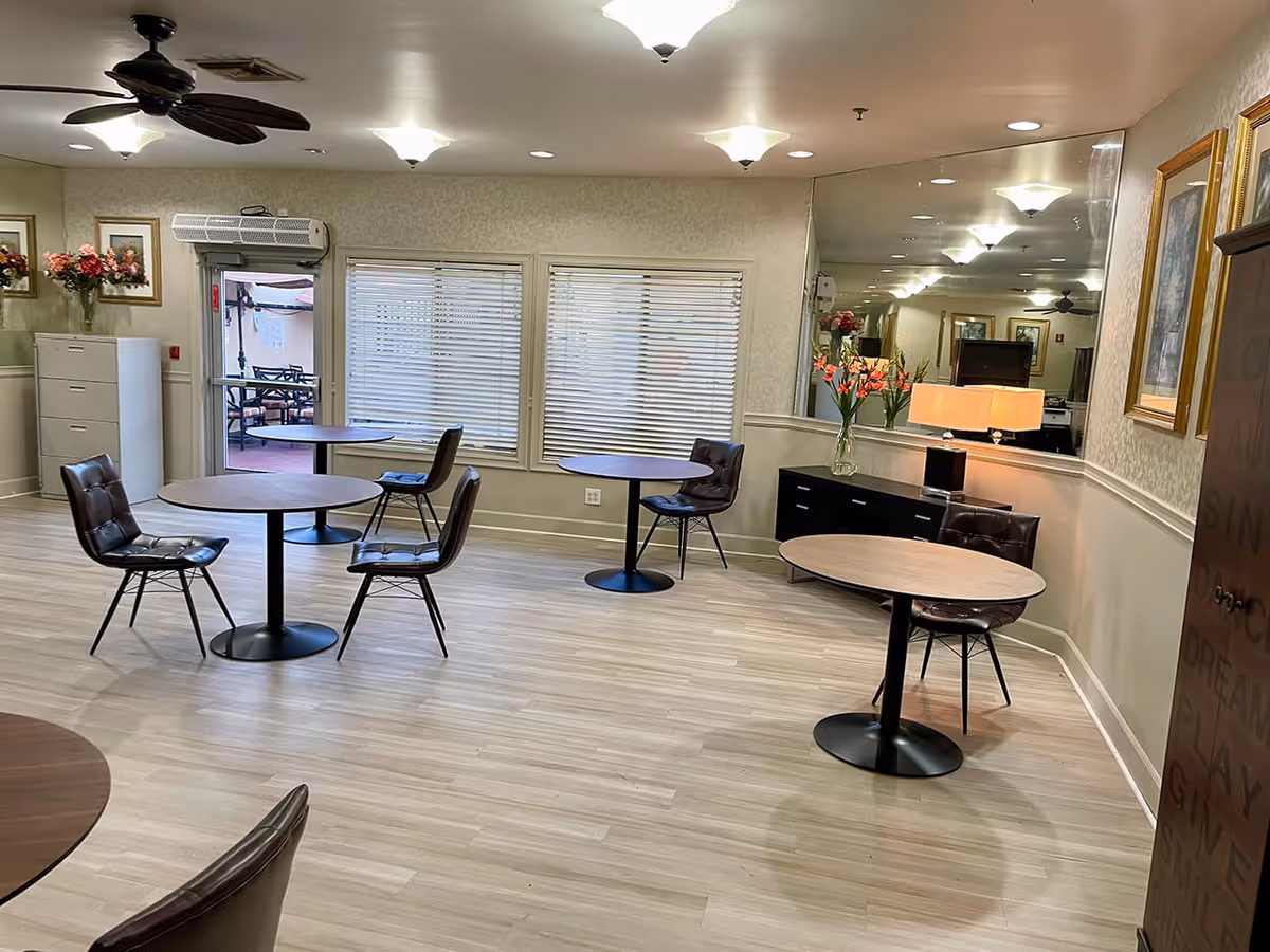 Bright communal dining room with round tables and four-legged chairs, floral arrangements, and a mirrored wall.