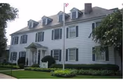 Large white two-story building with multiple windows and dormers on the roof, a small entrance porch, a flagpole with a flag in front, and well-maintained green lawn and bushes surrounding the building.