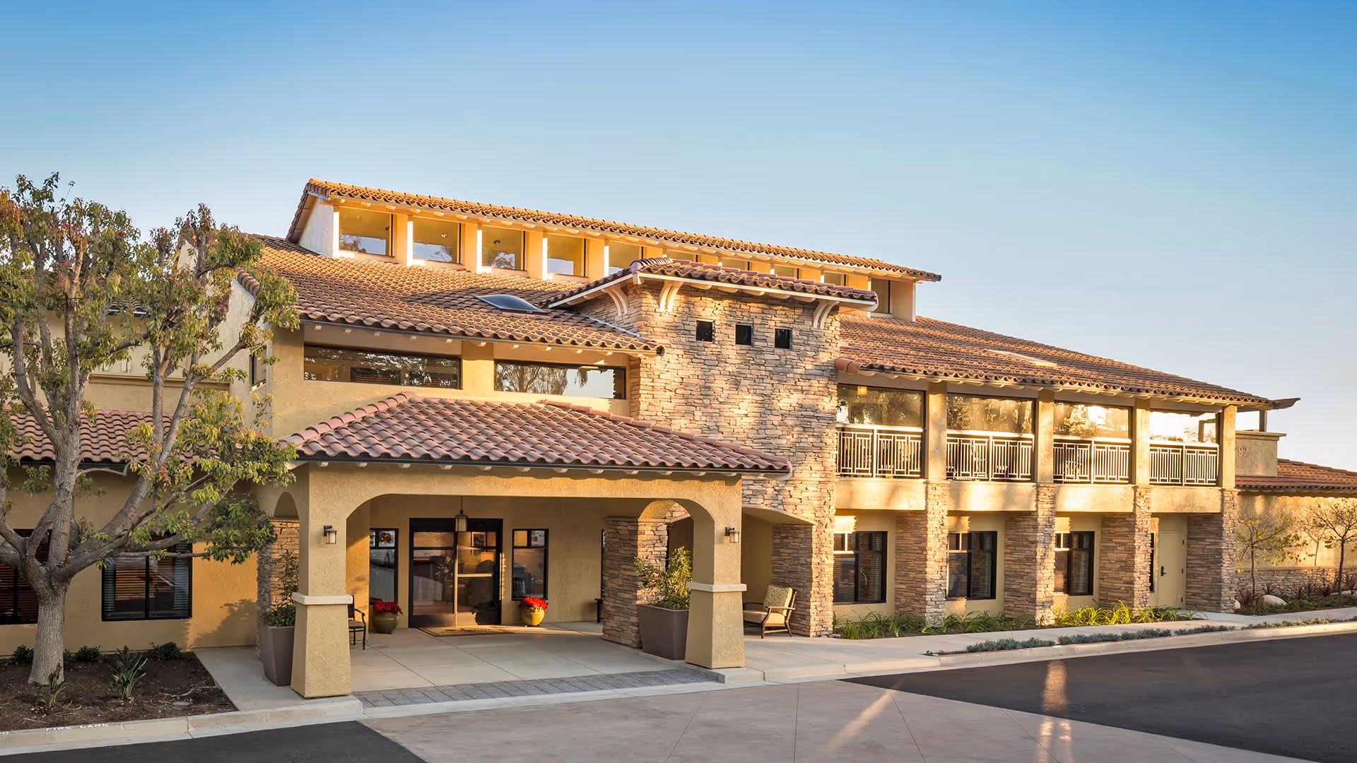Exterior view of Atria Del Sol, a two-story building with a tiled roof, stone and stucco facade, large windows, and a covered entrance with potted plants and seating areas.