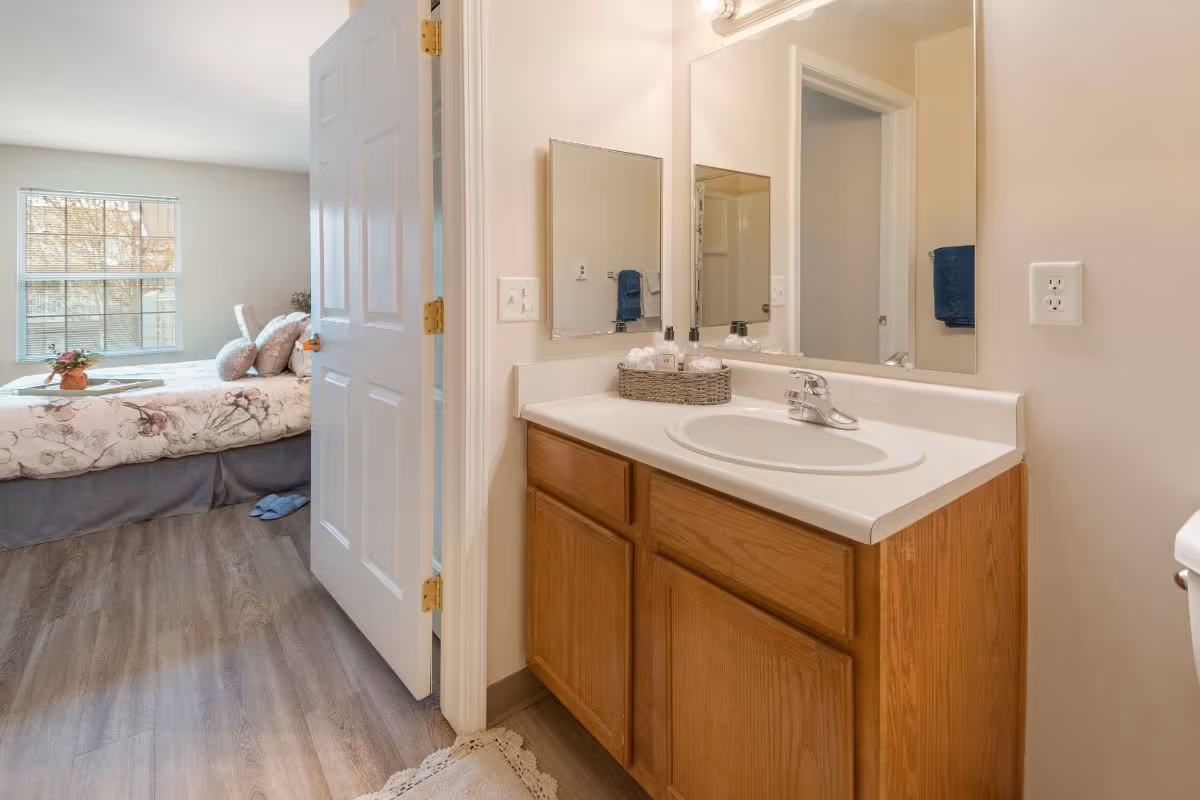 View of a bathroom sink with a wooden cabinet and a large mirror above it, next to an open door leading to a bedroom with a bed, pillows, and a window letting in natural light.