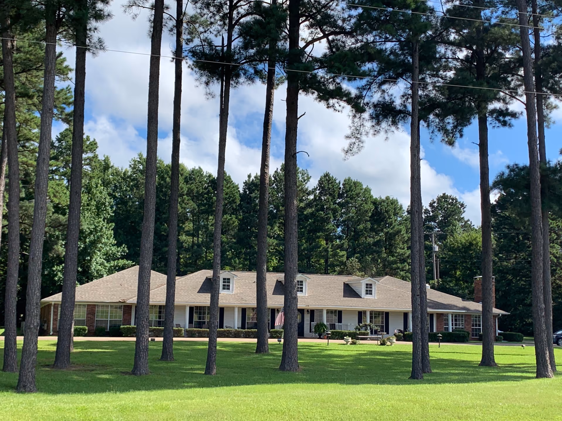 Single-story retirement lodge with a manicured lawn and tall pine trees in front.