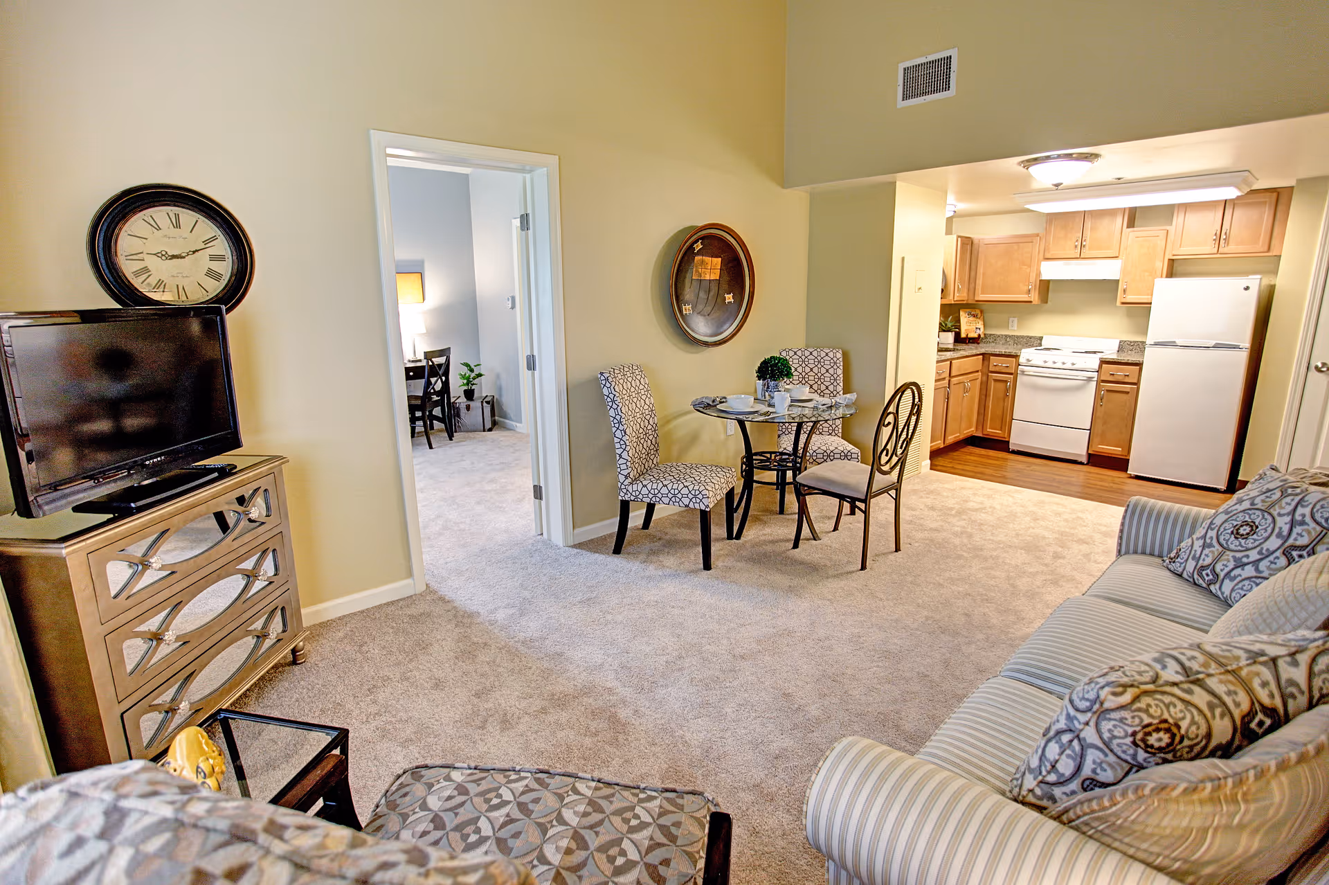 Interior view of a senior living facility apartment showing a living room with a striped sofa and patterned cushions, a small round dining table with three chairs, and an open kitchen with wooden cabinets, a stove, and a refrigerator. A doorway leads to another room with a desk and chair visible. A large wall clock is mounted above a TV on a mirrored dresser.