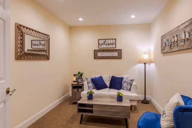 A cozy living room with beige walls and carpeted floor featuring a white loveseat with blue and patterned throw pillows, a wooden coffee table with small potted plants, a side table with a phone and plant, a floor lamp, and wall decorations including a mirror, a welcome sign, and a painting of birds on a branch.