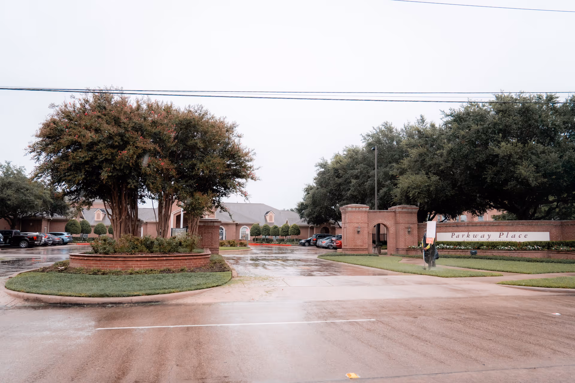 Brick entrance and driveway with a landscaped roundabout, trees, and parked cars at the Parkway Place facility on a rainy day.