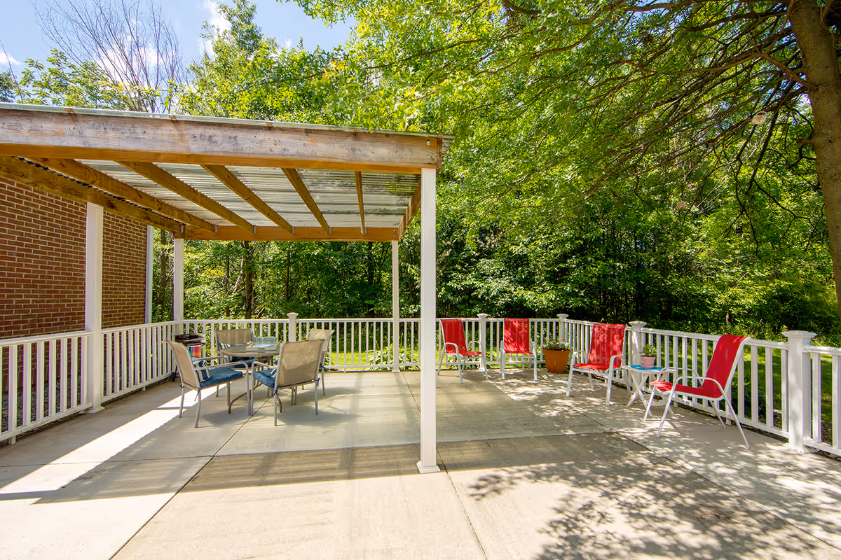 Outdoor patio area with a wooden pergola covering a table and chairs. Additional red and white chairs are arranged along a white railing. The patio is surrounded by lush green trees and grass under a sunny sky.