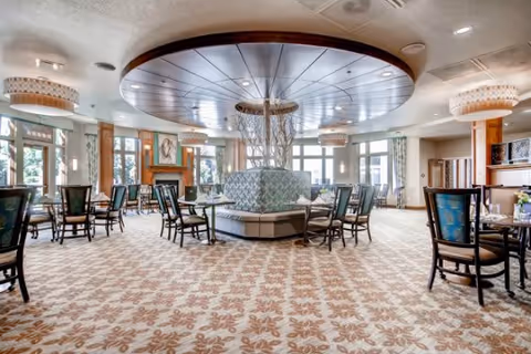 Spacious dining room with patterned carpet, multiple tables and chairs arranged neatly, large circular ceiling feature with decorative lighting, and large windows allowing natural light to fill the room.