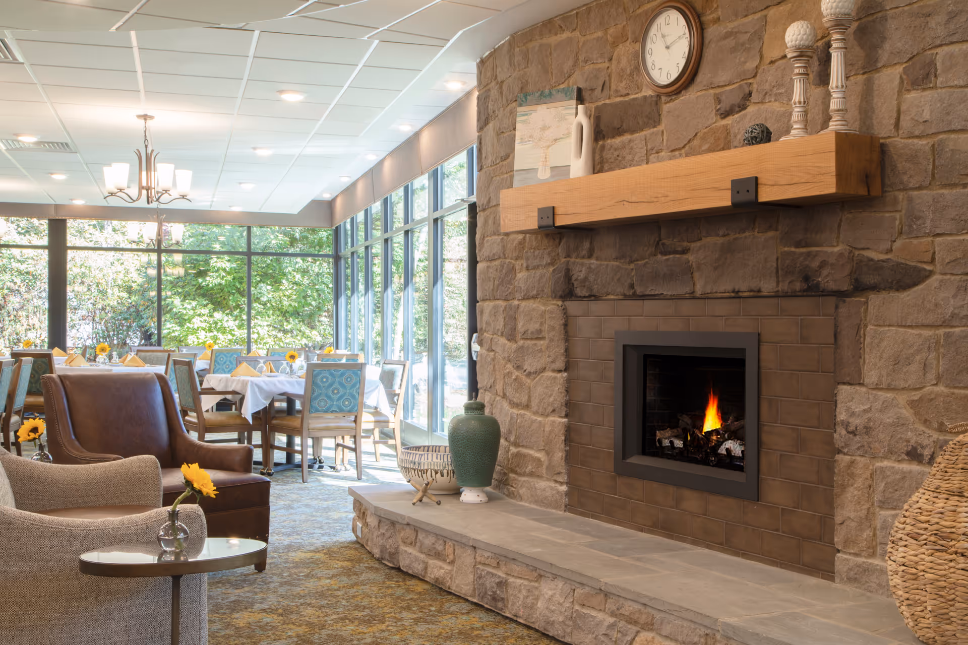A cozy dining and lounge area with large windows letting in natural light. The room features a stone fireplace with a wooden mantel decorated with vases and candlesticks. There are several tables with white tablecloths and chairs, some with blue patterned upholstery. Comfortable armchairs and a small round table with a single yellow flower in a vase are in the foreground. Outside, greenery is visible through the windows.