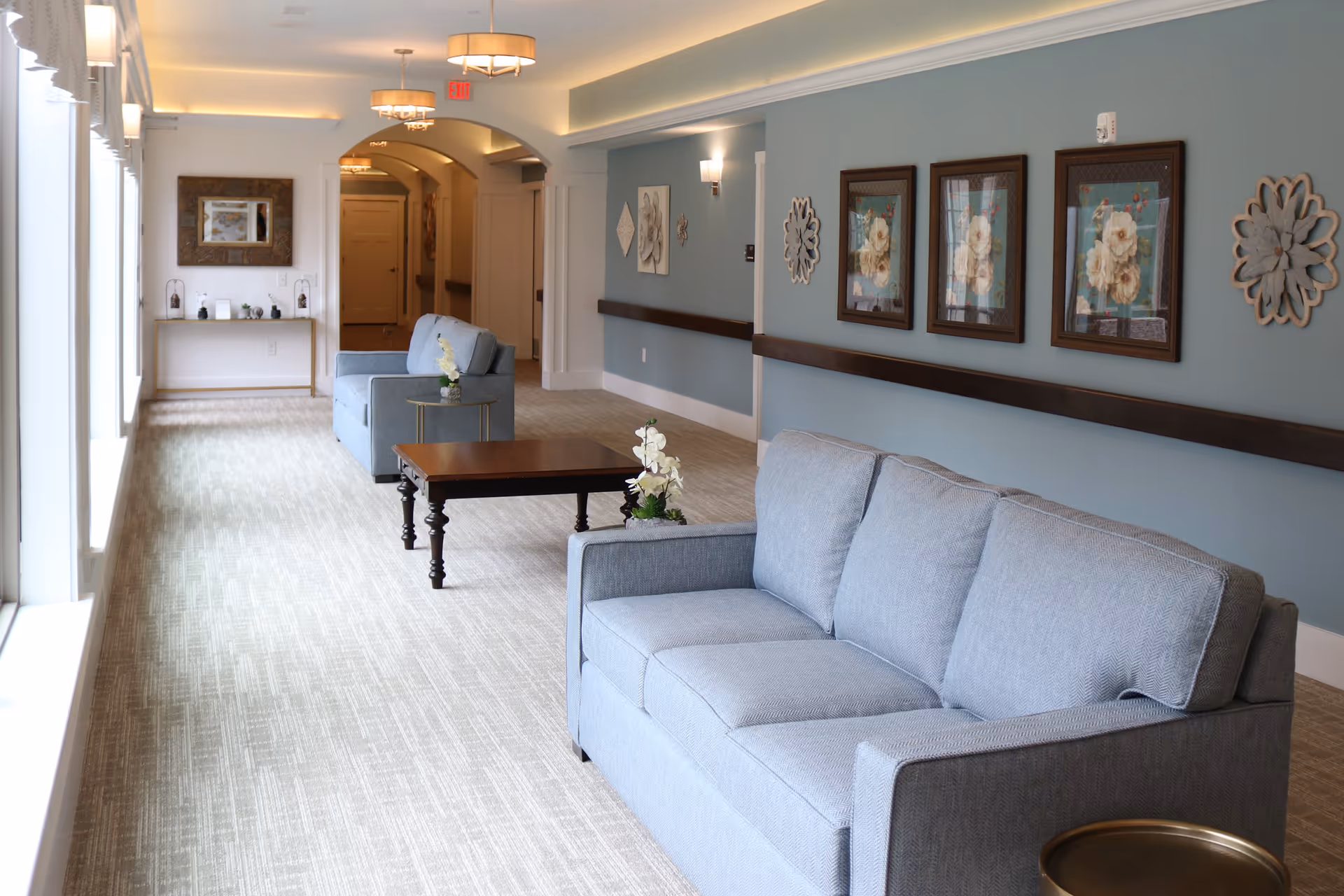 A bright and spacious hallway in a senior living facility with light blue walls and beige carpet. The hallway features two light blue sofas facing each other with a wooden coffee table in between. Decorative floral artwork and wall sconces adorn the walls. Large windows on the left side allow natural light to fill the space.