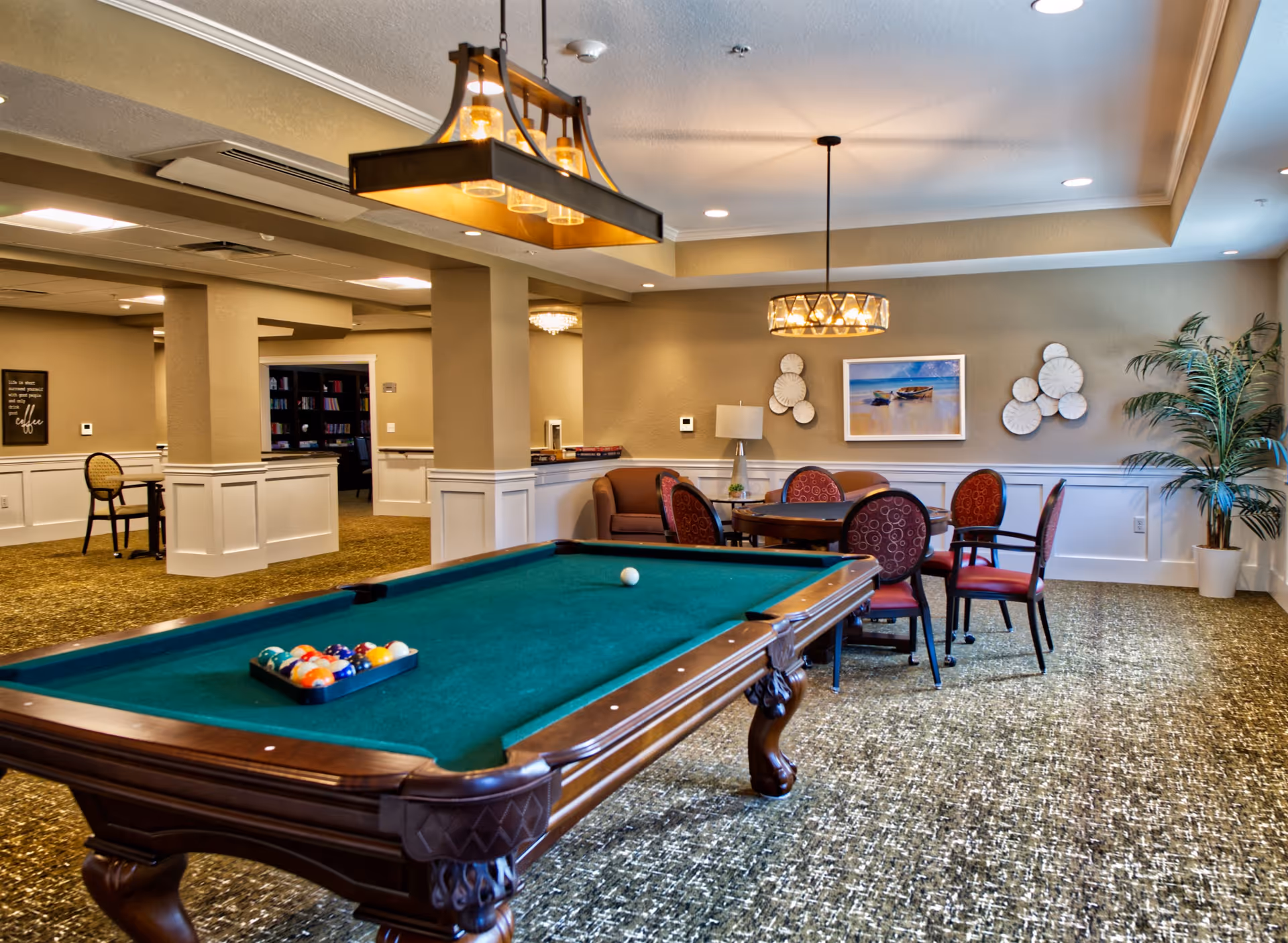A well-lit recreational room featuring a green felt pool table with billiard balls arranged for a game. Behind the pool table, there is a round table with four red cushioned chairs and a small sofa against the wall. The walls are decorated with a framed beach scene painting and circular wall art. The room has beige walls with white wainscoting, carpeted floor, and two hanging light fixtures. A potted plant is placed in the corner near a window.