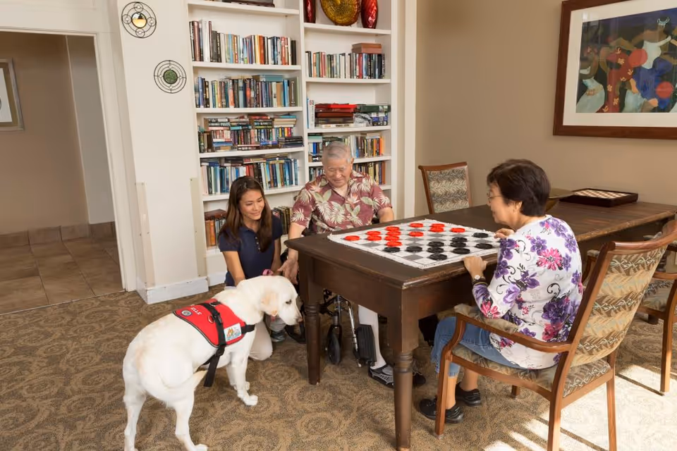 Three people in a cozy room with bookshelves playing a large checkers game on a wooden table. One person is seated in a wheelchair, another is sitting on a chair, and a third person is kneeling on the floor petting a white dog wearing a red service vest. The room has carpeted flooring, a framed colorful artwork on the wall, and a doorway leading to another area.