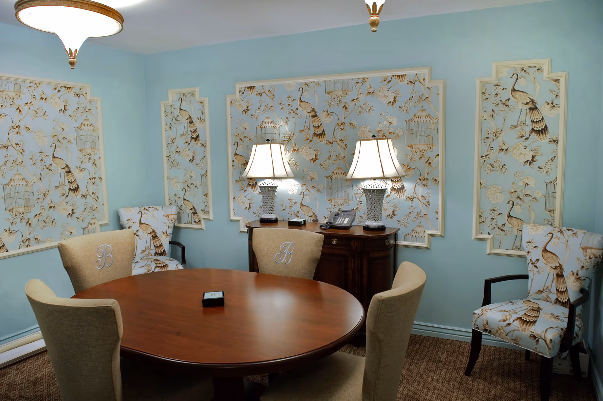 A small meeting or dining room with a round wooden table surrounded by four beige upholstered chairs, two of which have the letter 'B' embroidered on the back. The walls are light blue with decorative panels featuring a floral and bird pattern in beige and brown tones. A wooden sideboard with two white table lamps and a telephone sits against the wall. The room is carpeted and softly lit by ceiling lights.