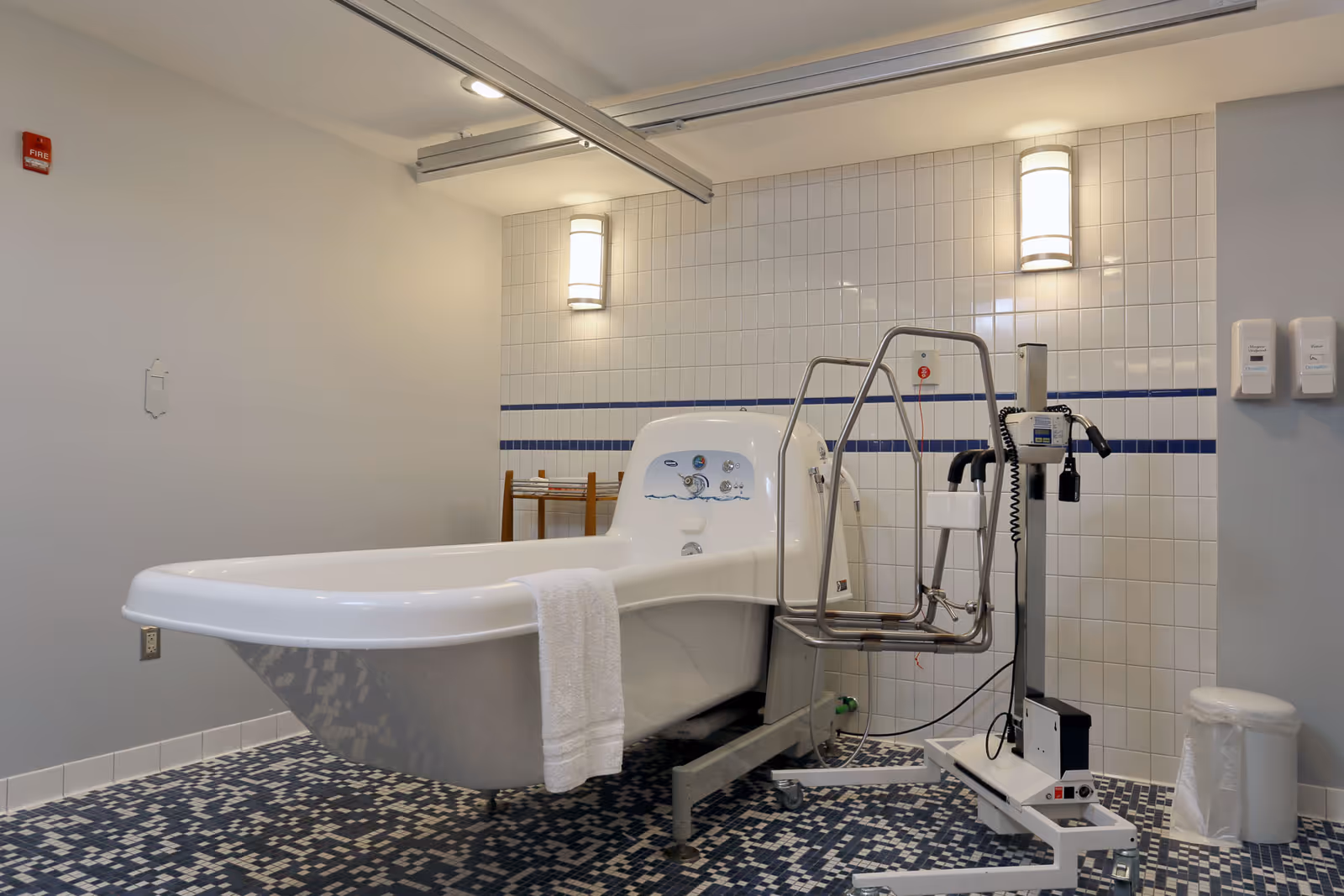 A clinical bathroom with a large white medical bathtub equipped with handrails and control knobs. The room has white tiled walls with a blue stripe and mosaic tile flooring in shades of blue and white. There are two wall-mounted lights above the tub, a small wooden rack in the corner, a white trash bin, and two wall-mounted dispensers on the right side.