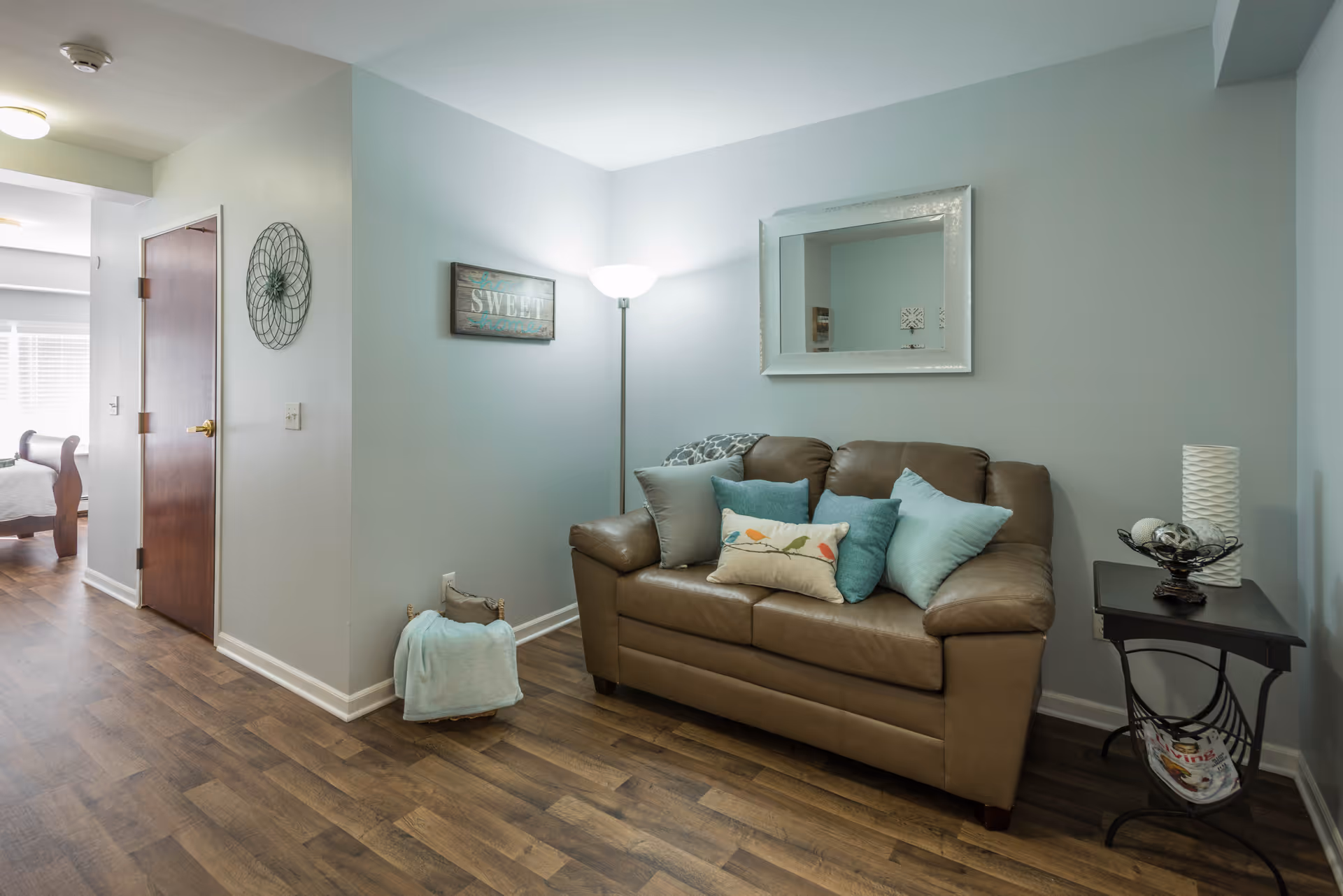 A cozy living room area with a brown leather loveseat adorned with multiple decorative pillows. A floor lamp is positioned in the corner behind the loveseat, illuminating the space. A rectangular mirror hangs on the wall above the loveseat, and a small side table with decorative items and a magazine rack is placed to the right. The room has light blue walls and wood flooring. A hallway leads to a bedroom with a wooden bed frame visible in the background.