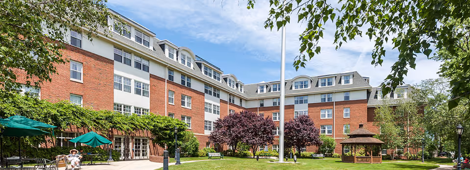 Exterior view of a multi-story brick assisted living facility with white window frames and a gray roof. The building surrounds a well-maintained courtyard with green grass, trees, benches, a gazebo, and outdoor seating with green umbrellas. A flagpole stands in the center of the courtyard under a partly cloudy sky.