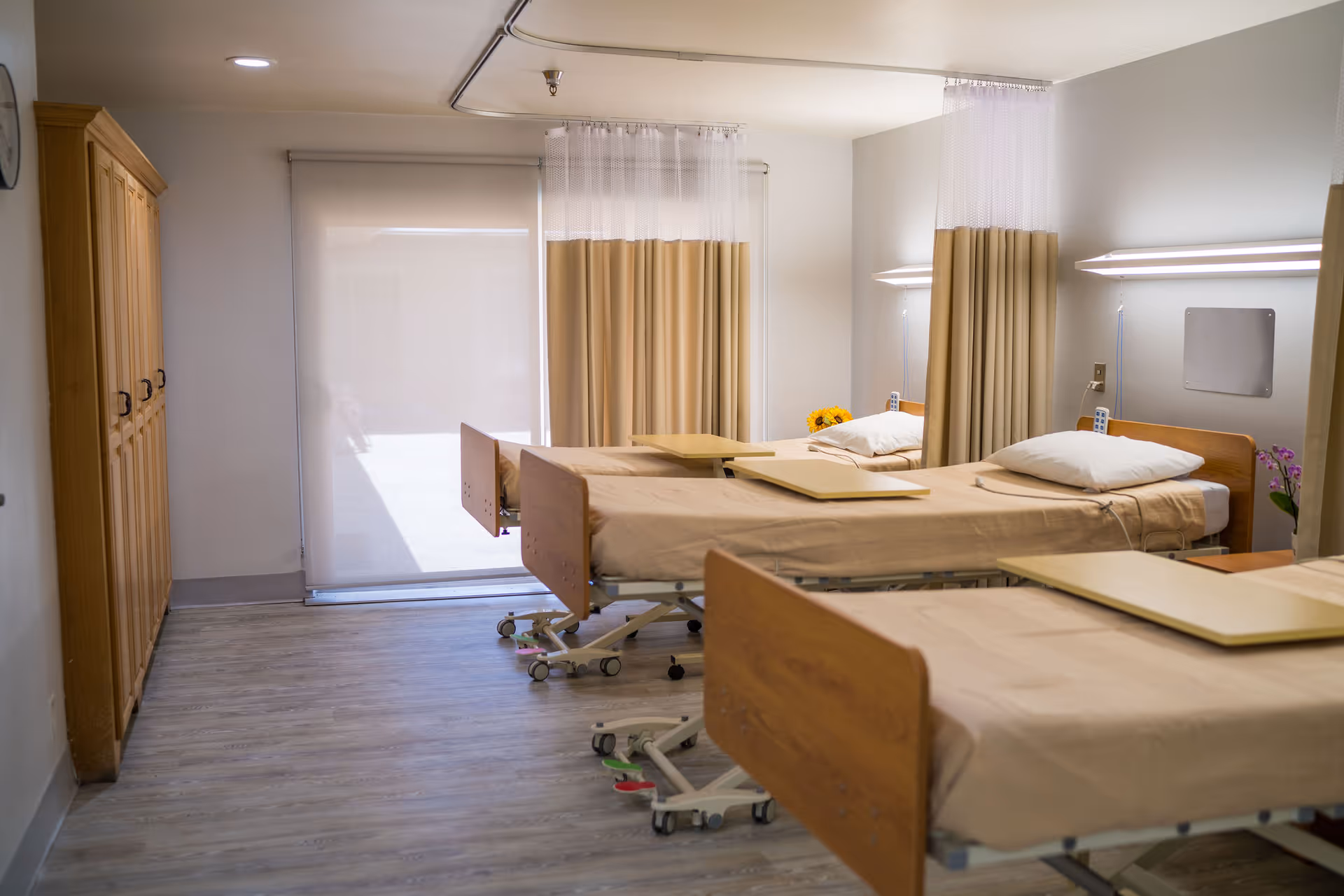 A hospital room with three beds arranged in a row, each bed equipped with beige sheets, pillows, and overbed tables. Privacy curtains hang between the beds, and there are wall-mounted lights above each bed. A wooden wardrobe is visible on the left side of the room, and a large window with a roller shade is at the far end.