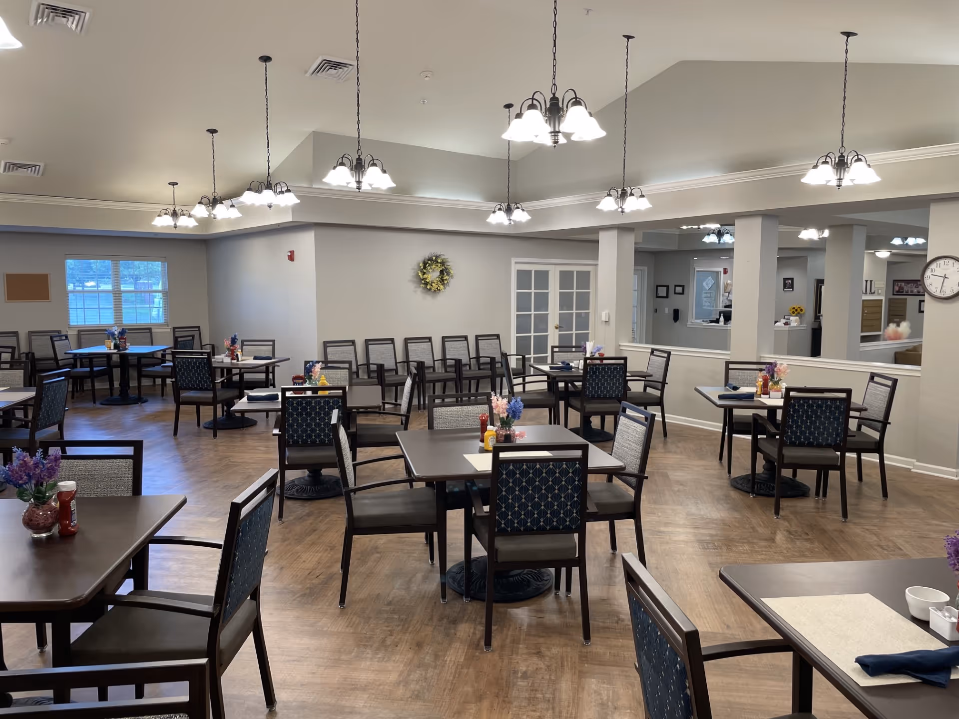 A spacious dining room in Cedar Creek of Bedford with multiple square and round tables, each set with placemats, napkins, and small flower arrangements. The room has wooden flooring, neutral-colored walls, and several hanging light fixtures providing bright illumination. There are windows on one side and a clock on the wall near an open reception area.