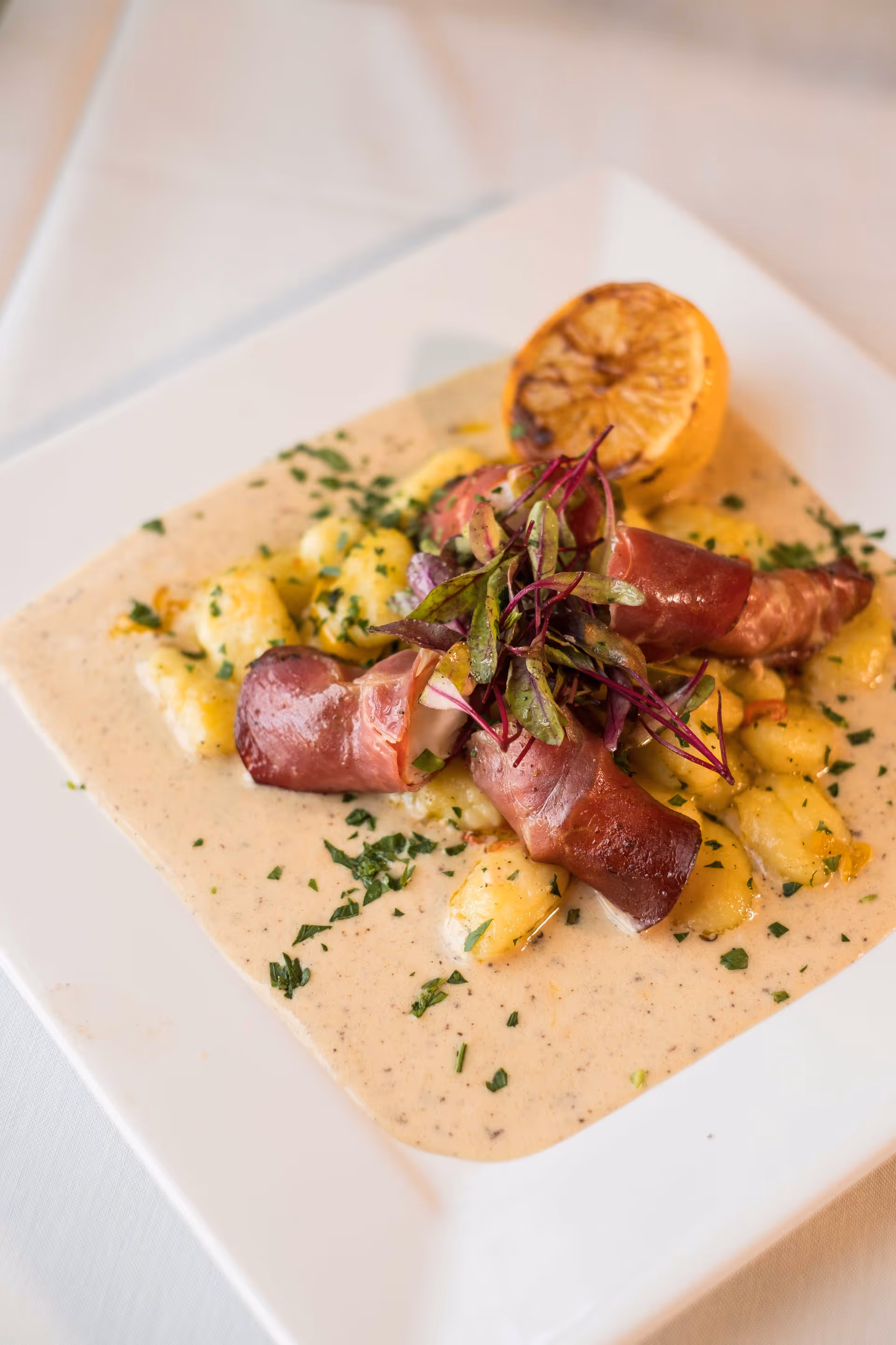 A gourmet dish featuring gnocchi in a creamy sauce, topped with rolled slices of cured meat and garnished with fresh herbs and microgreens, served on a white square plate with a grilled lemon half.
