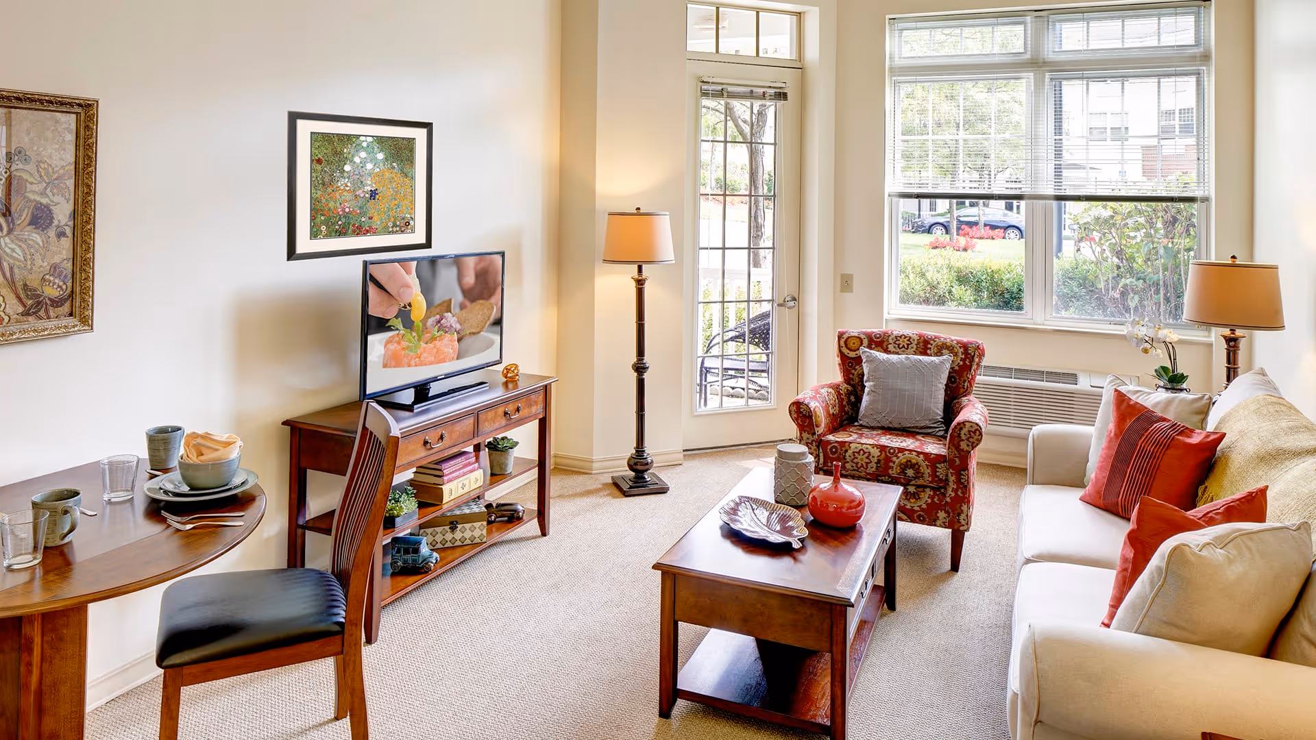 A bright and cozy living room with a beige sofa adorned with red and beige pillows, a patterned armchair with a gray pillow, a wooden coffee table with decorative items, a wooden TV stand with a flat-screen TV showing a close-up of food preparation, a small dining table with two place settings, and large windows letting in natural light.