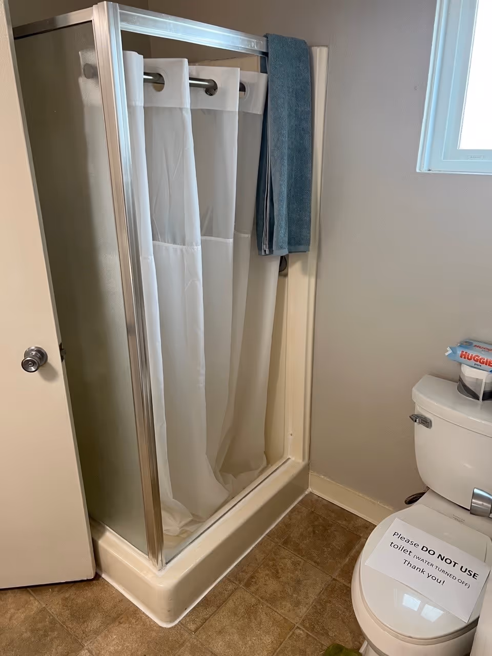 Compact bathroom showing a corner shower with a white curtain and towel hanging, and a toilet with a 'Do Not Use' sign on the seat.