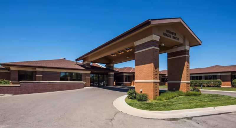 Exterior view of Brewster Health Center showing the main entrance with a covered driveway supported by brick pillars, surrounded by a paved driveway and landscaped greenery under a clear blue sky.