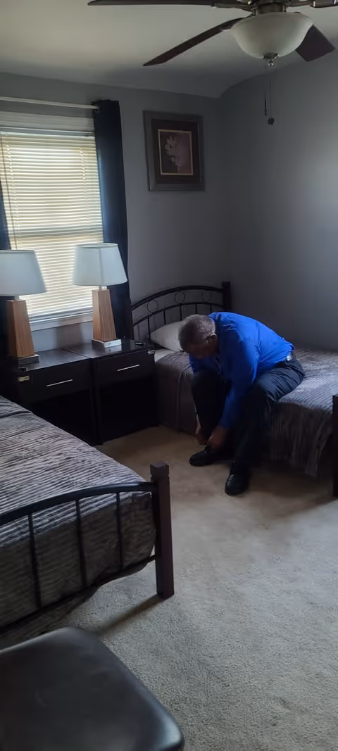 A man in a blue shirt sitting on a bed in a bedroom, bending down to tie his shoe. The room has two single beds with gray bedspreads, a nightstand with two lamps between the beds, a window with blinds and dark curtains, a ceiling fan, and a framed picture on the wall.