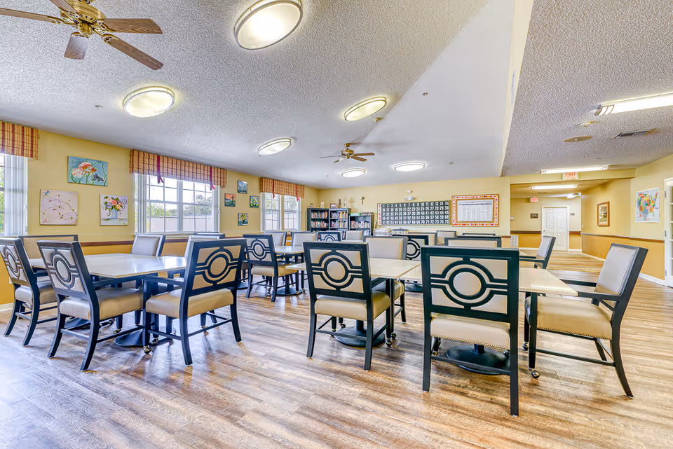 Spacious senior dining/activity room with multiple tables and chairs, large windows, ceiling fans, and wall decor.