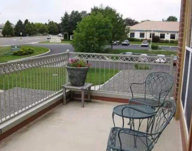 A small outdoor balcony or patio area with two metal chairs and a matching small table. There is a potted plant on a small wooden stand near the railing. The balcony overlooks a parking lot, a grassy area, and a building in the background under a cloudy sky.
