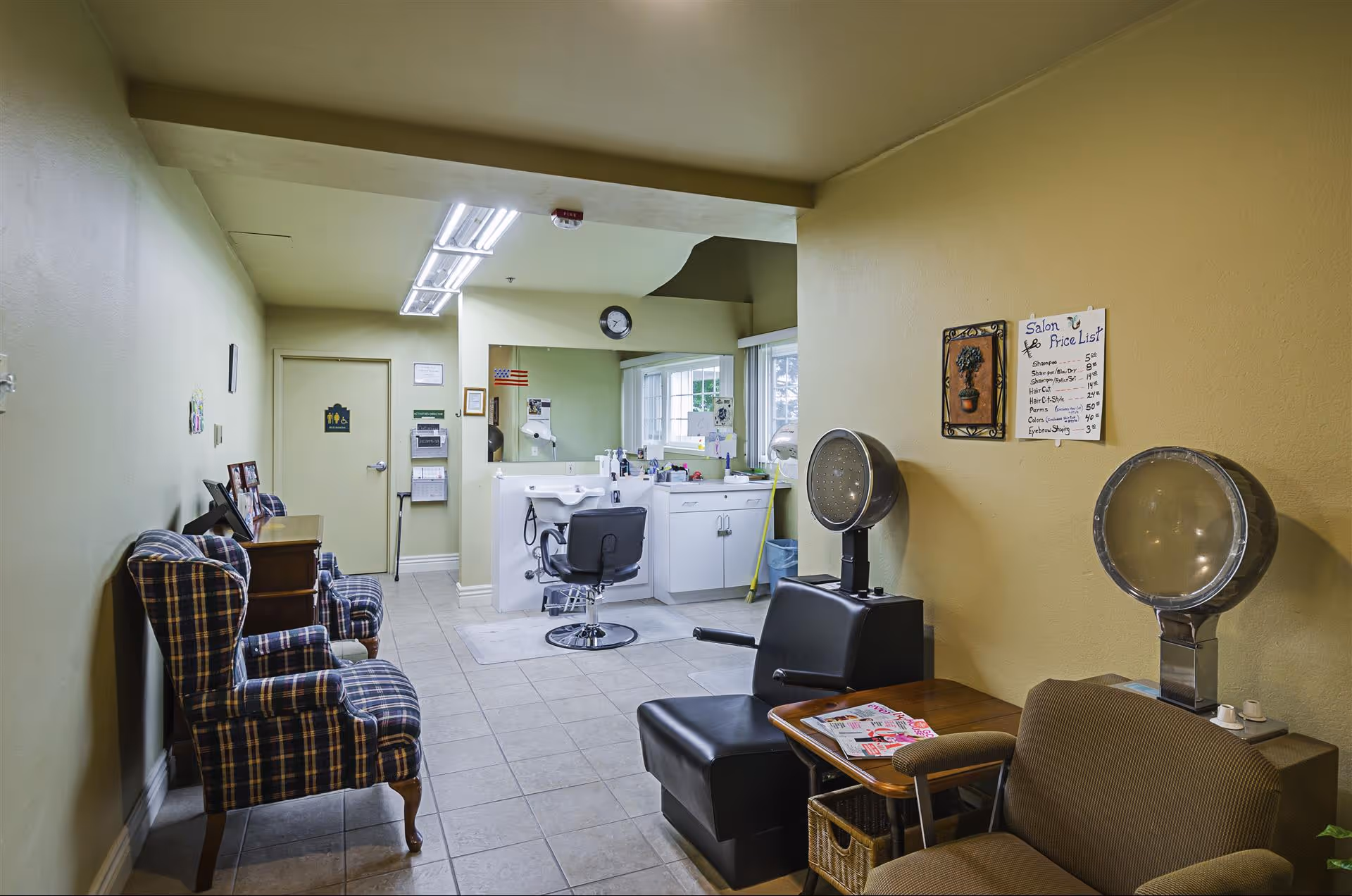 Interior view of a salon area in Cambridge Court Apartments featuring salon chairs, hair dryers, a sink, and a price list on the wall. There are also plaid upholstered chairs along the left wall and a small table with magazines.
