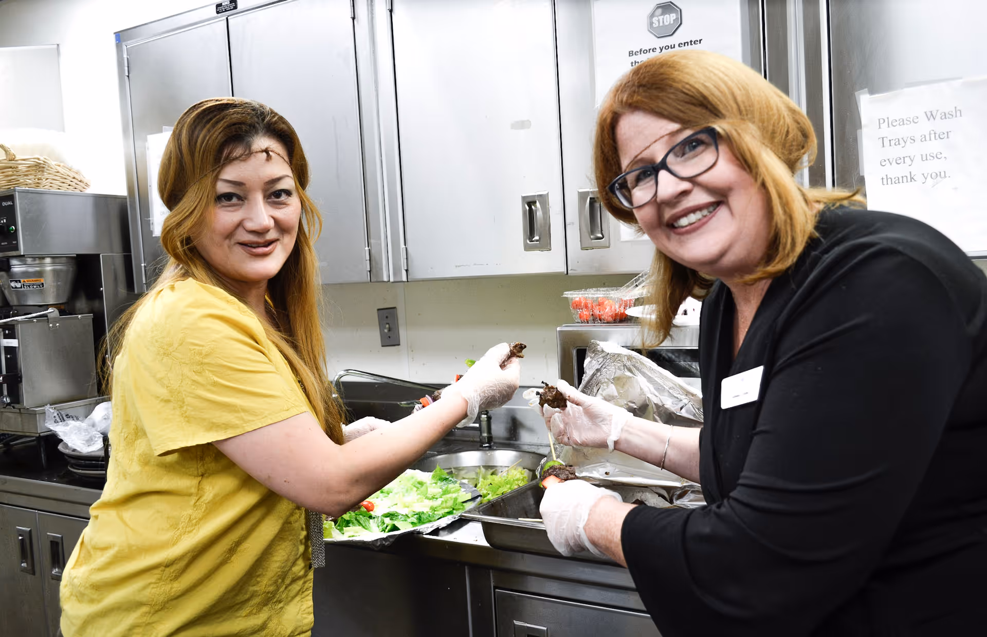 Two women wearing gloves preparing food in a kitchen. One woman is wearing a yellow shirt and the other is wearing a black top with glasses. They are smiling at the camera while handling food over a tray of lettuce and other ingredients in a stainless steel kitchen area.