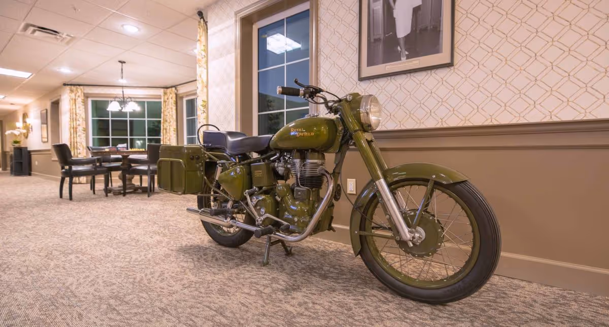 An olive-green Royal Enfield motorcycle displayed in a carpeted interior common area with a dining table in the background.