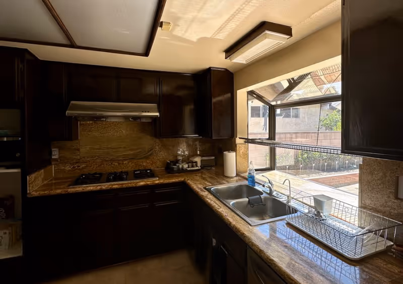 A kitchen with dark wooden cabinets, a gas stove with a range hood, a double sink, and a dish drying rack. There is a large window above the sink letting in natural light and showing an outdoor garden area.