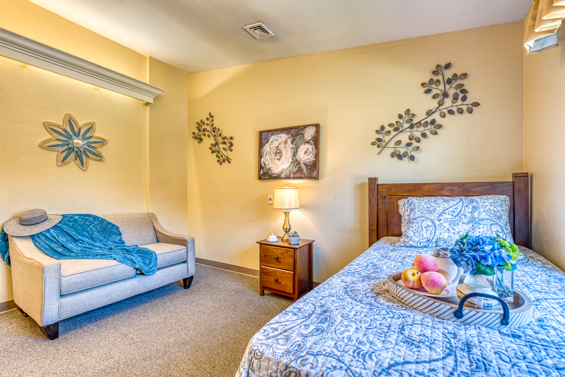 A cozy bedroom with a single wooden bed covered in a blue and white patterned quilt. On the bed is a tray holding peaches, a small vase with blue flowers, and a white pitcher. Next to the bed is a wooden nightstand with a lamp, a small plant, and a white device. A beige loveseat with a blue throw blanket and a hat is positioned against the opposite wall. The walls are decorated with floral metal art and a framed painting of white flowers. The room has beige walls and carpeted flooring.