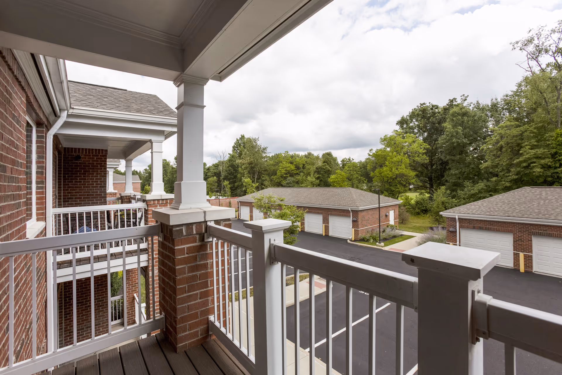 View from a second-floor balcony with white railings and brick pillars overlooking a parking area with garages and green trees in the background under a cloudy sky.