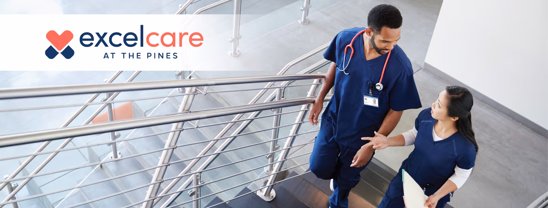 Two healthcare professionals in navy blue scrubs walking up a staircase inside a modern building, engaged in conversation. One has a stethoscope around his neck and the other is holding a clipboard. The Excelcare at the Pines logo is visible in the upper left corner.