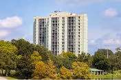 Tall white apartment building behind a tree-filled foreground under a blue sky.
