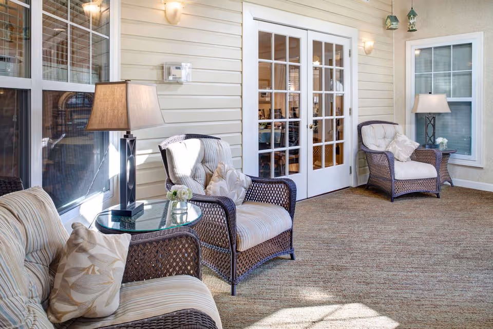 A cozy indoor seating area with three cushioned wicker chairs arranged around two glass-top side tables, each with a lamp and a small flower vase. The space features beige walls with white trim, two large windows, and a set of white French doors leading to another room.