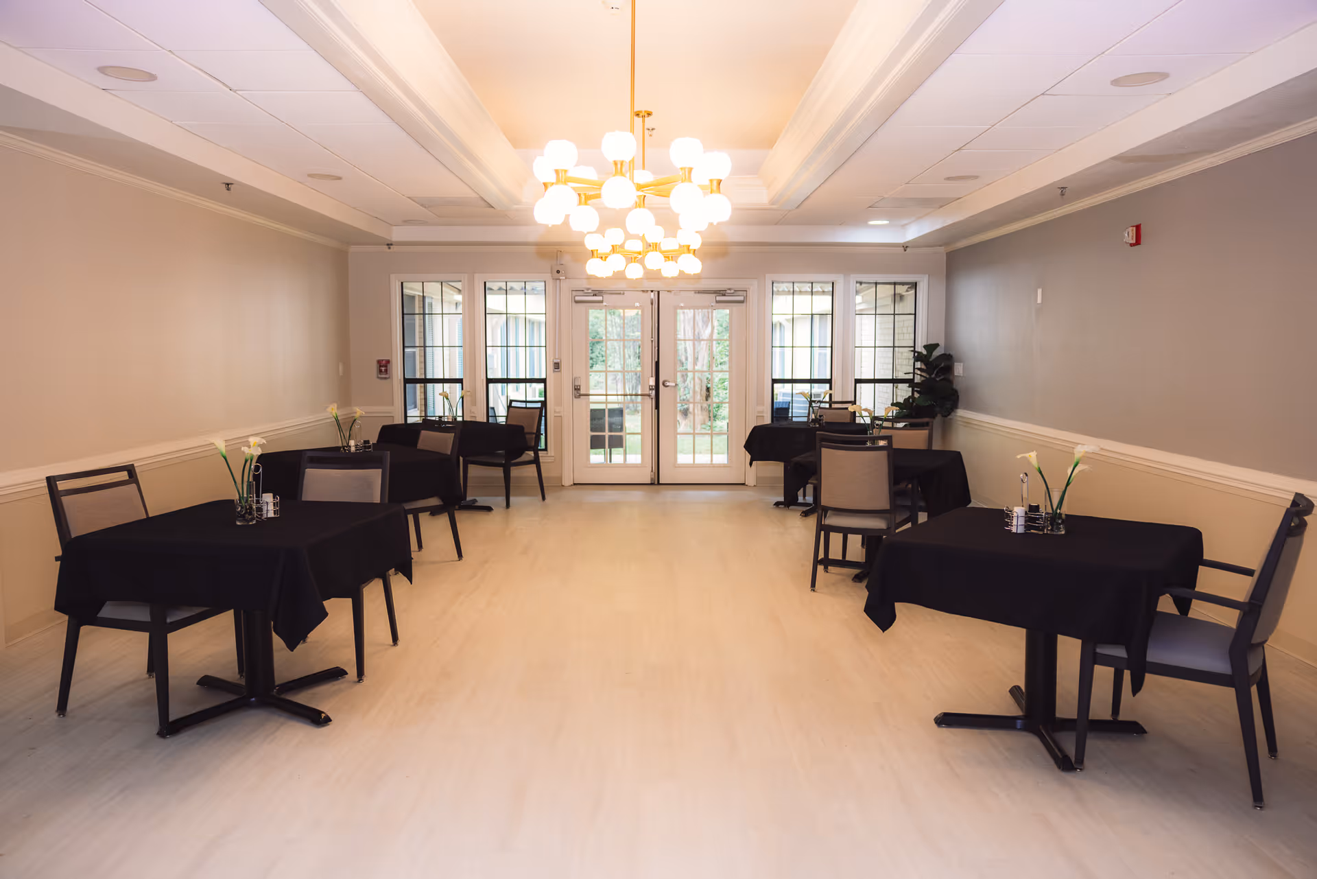 A dining room with several tables covered in black tablecloths, each set with chairs and small flower arrangements. The room has light-colored walls, a light wood floor, and a modern chandelier hanging from the ceiling. Large windows and glass doors at the far end let in natural light and show greenery outside.