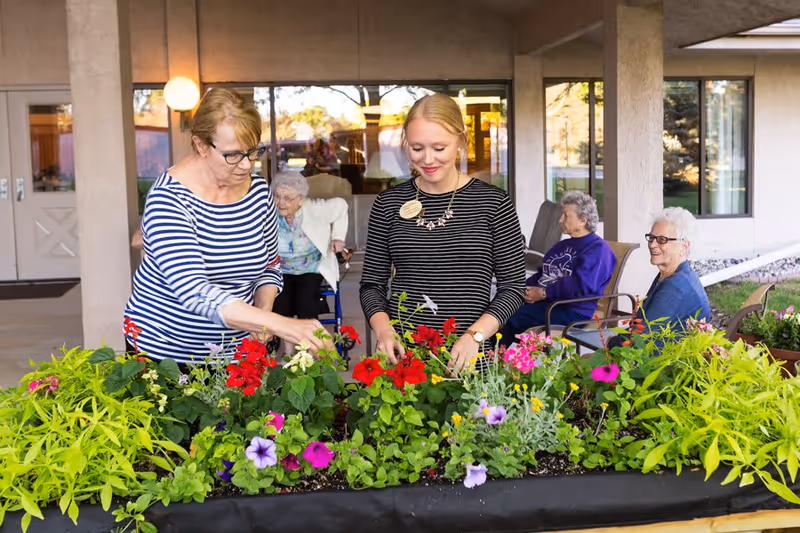 Two women tending to a raised flower bed filled with colorful flowers and greenery outside a building, with three elderly women sitting and observing in the background.