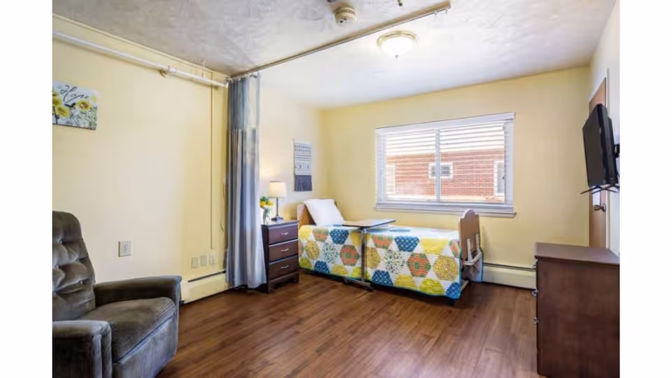 A bright and clean bedroom in a senior living facility with a single bed covered in a colorful quilt, a bedside table with a lamp and flowers, a comfortable armchair, a window with blinds, and a wall-mounted television.