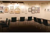 An interior room with beige walls displaying various framed artworks arranged in a gallery style. Several black chairs are arranged in rows facing the artwork on the walls. The room has a carpeted floor and ceiling lights illuminating the art pieces.