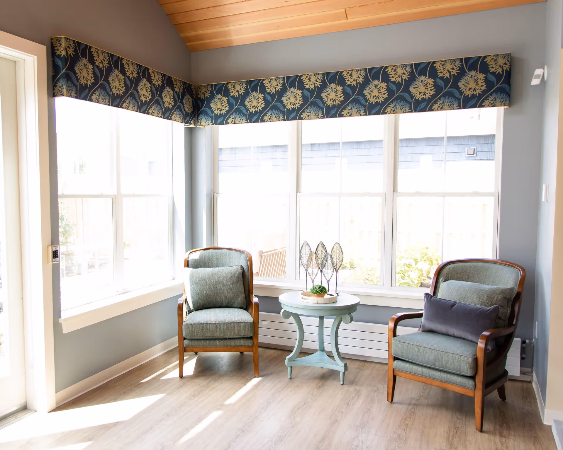 A bright corner seating area with two cushioned wooden armchairs and a small round table between them, decorated with a small plant and metal leaf sculptures. Large windows with floral valances allow natural light to fill the space, and the room has light wood flooring and soft gray walls.