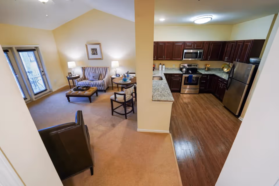 Interior view of a senior living facility apartment showing a living room and kitchen area. The living room has a striped loveseat, two side tables with lamps, a cushioned ottoman, a round dining table with two chairs, and a leather armchair. The kitchen features dark wood cabinets, granite countertops, stainless steel appliances including a refrigerator, stove, and microwave, and wood flooring. The walls are painted light yellow and there is a glass door leading outside.