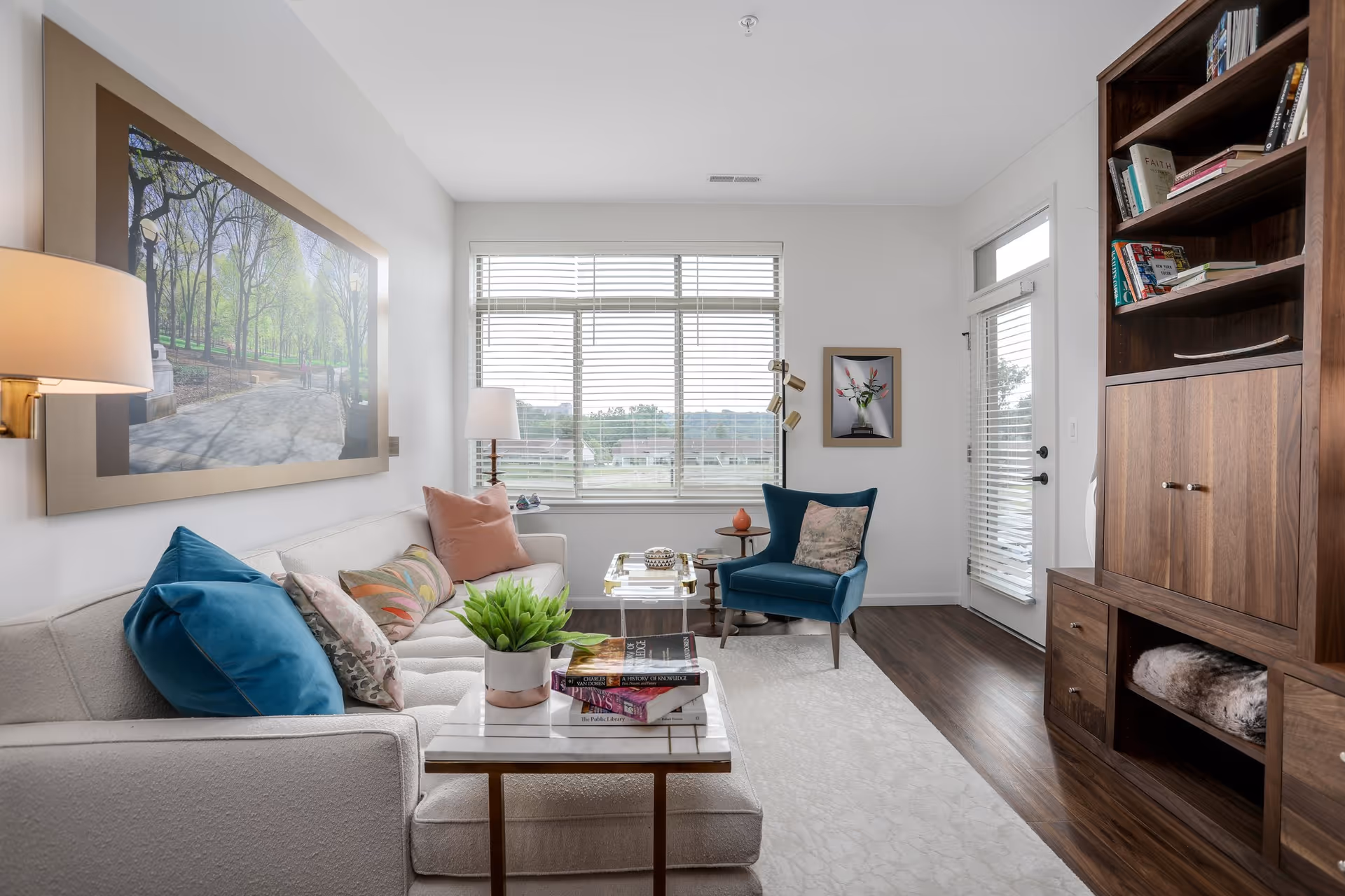A bright and cozy living room with a white sofa adorned with colorful pillows, a small marble-top side table with a potted plant and books, a blue armchair with a decorative pillow, a large window with blinds letting in natural light, a wooden bookshelf filled with books and decor, and framed artwork on the walls.