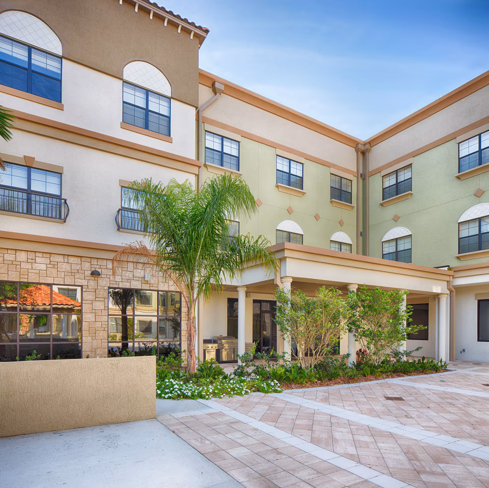 Exterior courtyard view of a multi-story senior living facility with beige, green, and tan walls, multiple windows, a covered patio area with columns, palm trees, and landscaped greenery under a clear blue sky.