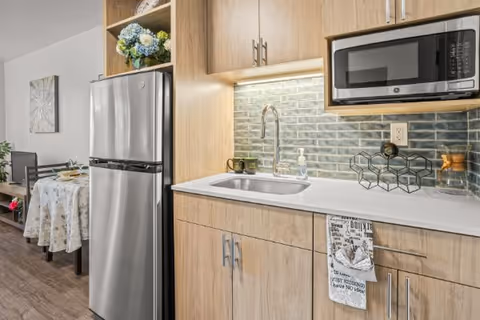 Modern kitchen area with light wood cabinets, a stainless steel refrigerator, a built-in microwave, a sink with a faucet, and a decorative backsplash. A small dining table with chairs and a floral tablecloth is visible in the background.