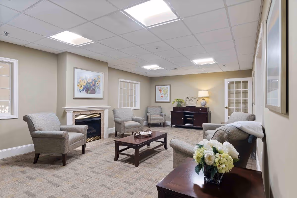 A cozy living room area with beige walls and carpeted floor. The room features four upholstered armchairs arranged around a wooden coffee table, a fireplace with a floral painting above it, a dark wooden sideboard with a lamp and decorative items, and a small table with a vase of white flowers in the foreground. The ceiling has recessed lighting and there are windows and a glass-paneled door on the walls.
