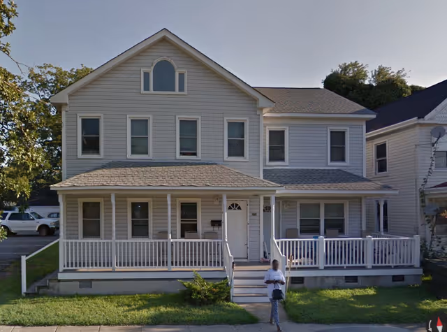 Front view of a two-story beige house with a wide covered porch, white railings, and a person standing on the walkway.