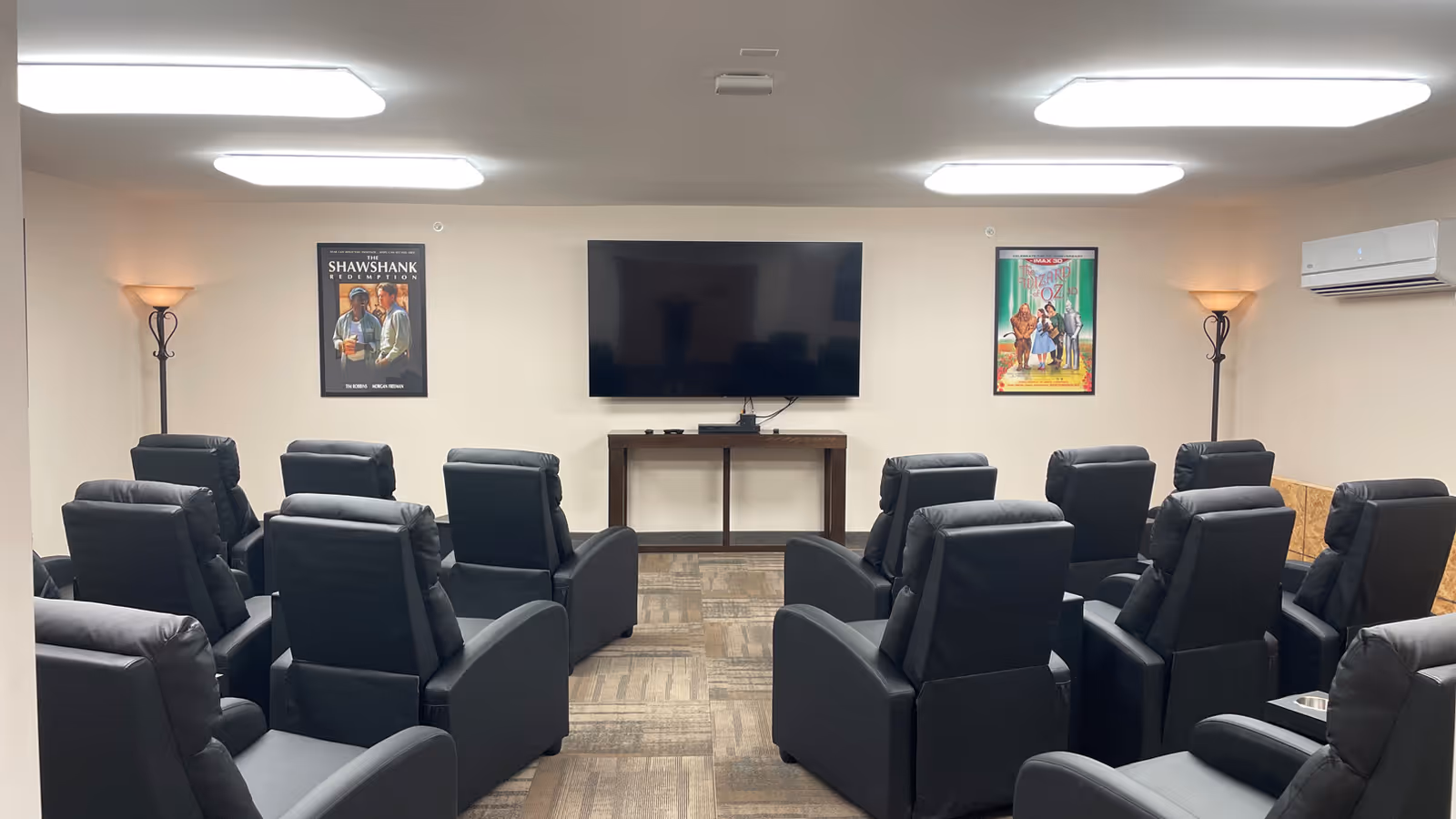 A small theater room with two rows of black recliner chairs facing a large flat-screen TV mounted on a beige wall. The room is lit by ceiling lights and two floor lamps in the corners. Movie posters for The Shawshank Redemption and The Wizard of Oz are hung on the wall beside the TV.