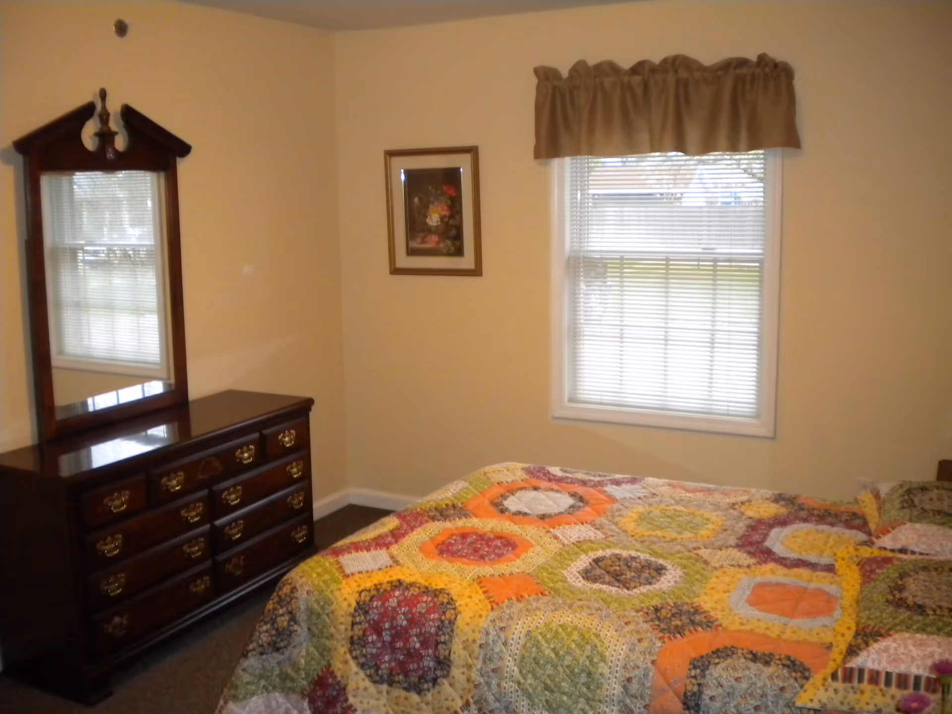 A simple bedroom with a colorful patchwork quilt on the bed, a wooden dresser with mirror, and a window with blinds and a valance.