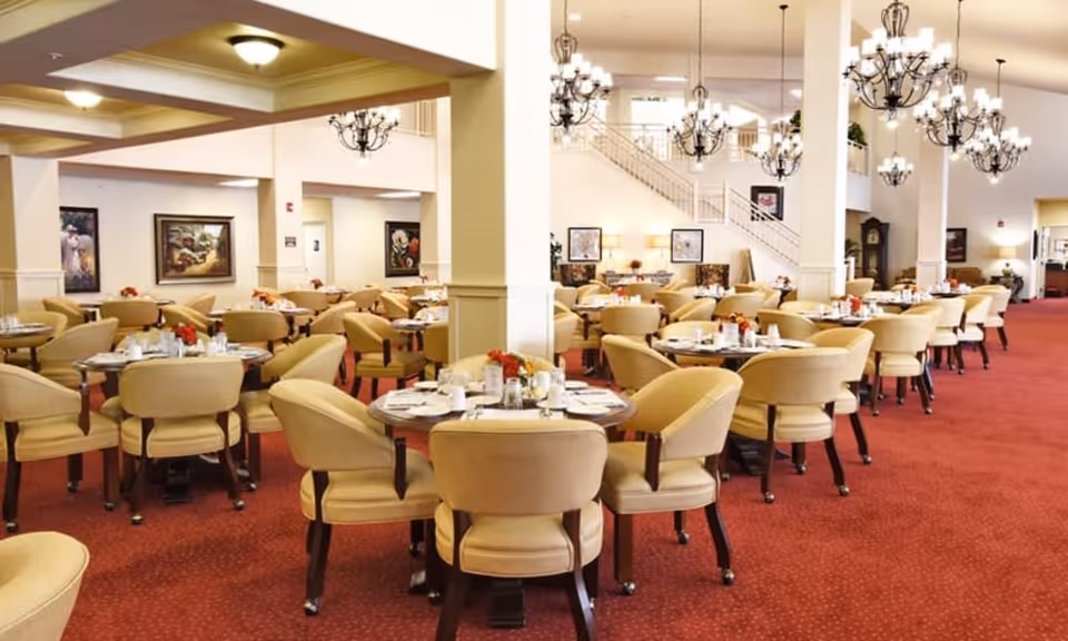 Spacious dining room with multiple round tables set with white plates, cups, and silverware. Each table is surrounded by beige upholstered chairs. The room features red carpet flooring, white walls with framed artwork, and several black chandeliers hanging from the ceiling. A staircase and additional seating areas are visible in the background.